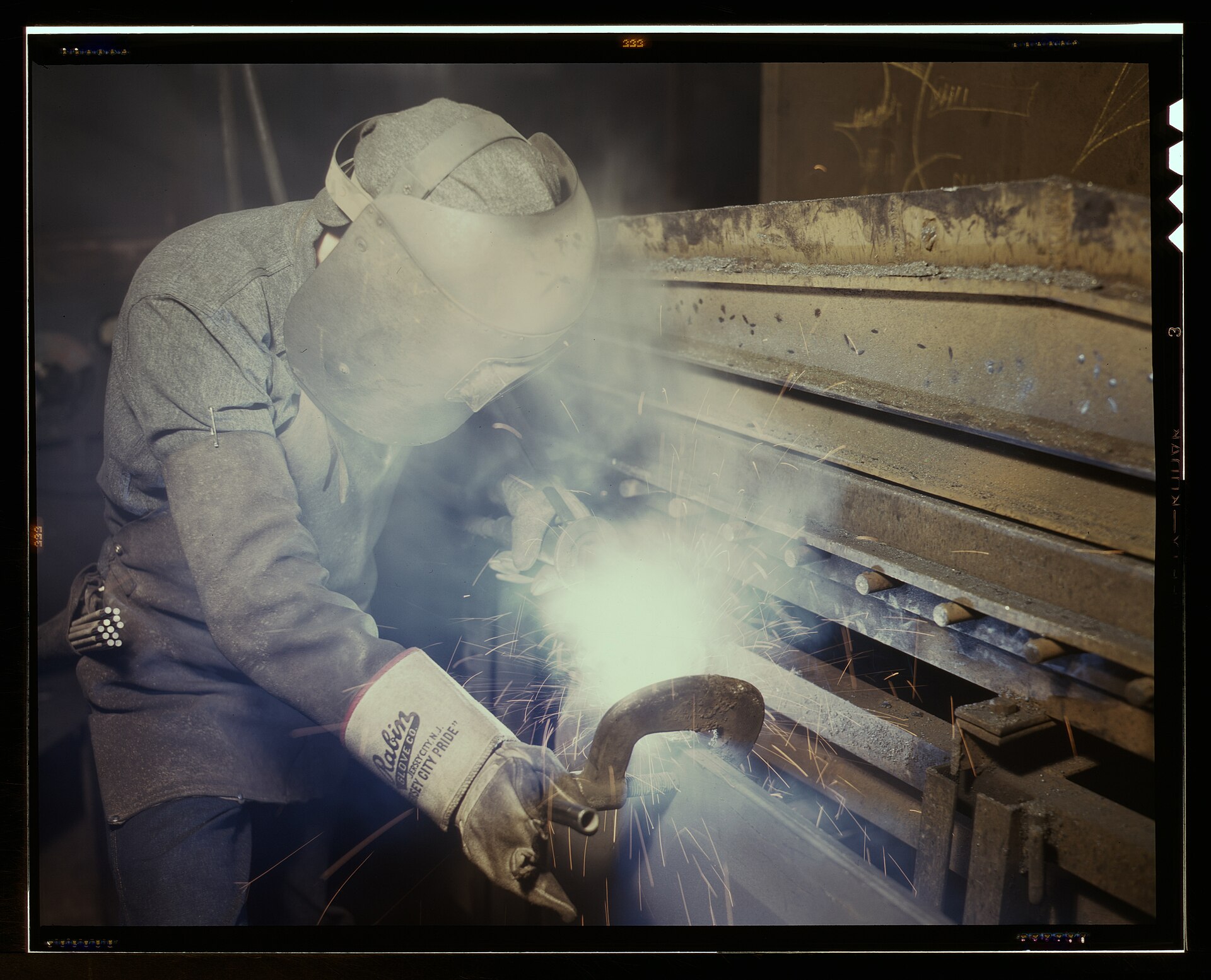 A welder in a mask and leather gloves welding a boiler seam in a shower of sparks.